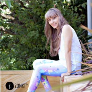A girl with long, wavy, light brown hair and light purple highlights is sitting on a bench outdoors. she is wearing a sleeveless white top and leggings in an abstract pastel print. she is seated side-on to the camera and is looking towards the photographer with a smile. The photo is watermarked with the Zonkt logo in the bottom left corner.