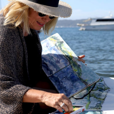 Ally Bryan pictured seated by the ocean painting in a sketchbook.