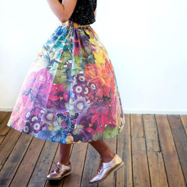 A cropped waist-down photo showing a person in a voluminous mid-length skirt made in a colourful floral design created by Anika Cook. The person is wearing fancy metallic pink shoes.