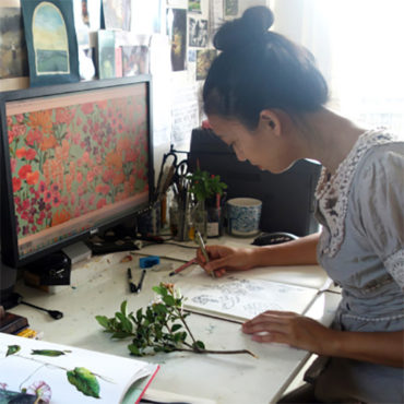 Cecilia Mok is seated at a desk drawing in a sketchbook, she is referencing floral illustrations and a fresh cut flower.