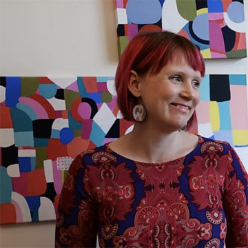 A portrait of Jiah Harrison of Elephant and Rose standing in front of a collection of her paintings. Jiah has chin length straight, bright red hair and is facing to the right. She is wearing a wide-neck long sleeve top with a red pattern and navy blue background.