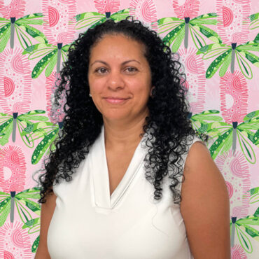 Ivy Helena is pictured in front of a fun pink and green Australian Native floral design. She has curly black hair and is wearing a white v-neck sleeveless top.