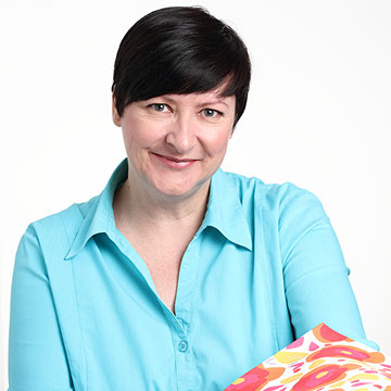 A portrait of Julie Harrison, a white woman with short dark hair wearing a turquoise button up blouse holding a folded piece of fabric showing her Collage Flower Orange Red print.