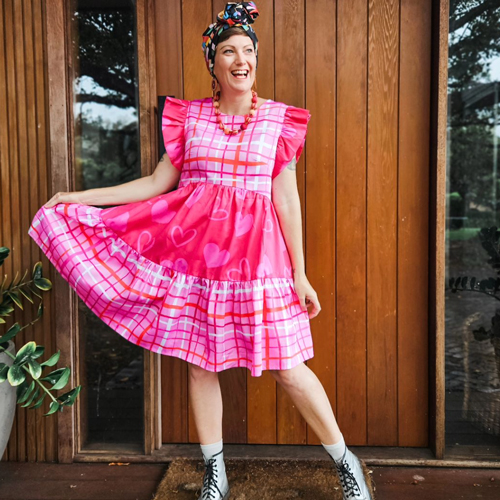 Kathryn Shaw of brand Rattamatatt wearing a bright pink dress standing in a. doorway smiling.