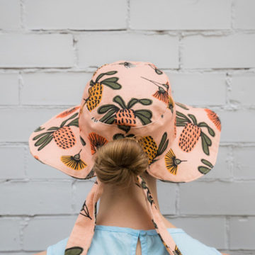 A person with strawberry blonde hair in a bun is standing with their back towards the camera in front of a sage green painted brick wall. They are wearing a robin's egg blue sleeveless shirt and a sunhat with a peach coloured banskia print designed by Indigo Thread.