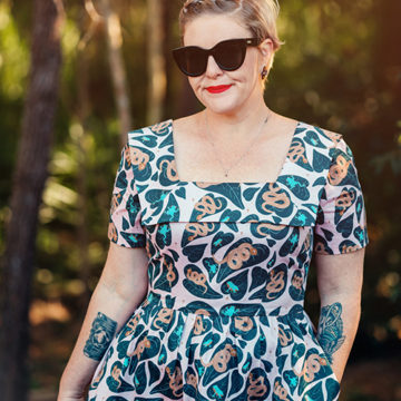 A woman wearing a dress made using a Katie Makes a Dress design and black sunglasses in a softly lit forest. The dress pattern, QLD Summer, is turquoise frogs and brown snakes on dark green leaves. The woman has red lipstick and black tattoos on her forearms.