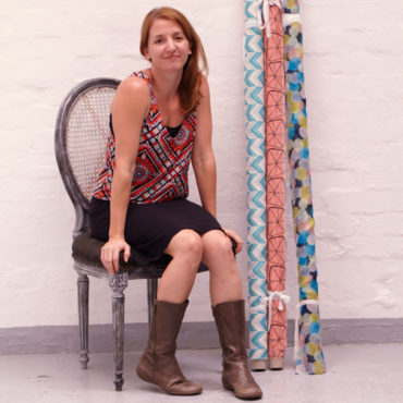 Lily Fink is shown seated on a vintage chair in front of a painted white brick wall. There is three rolls of fabric leaning against the wall to her right. Lily has long straight red hair and is wearing a black knee-length skirt, brown mid-calf leather boots, and a mulitcolour tank top with a geometric design.