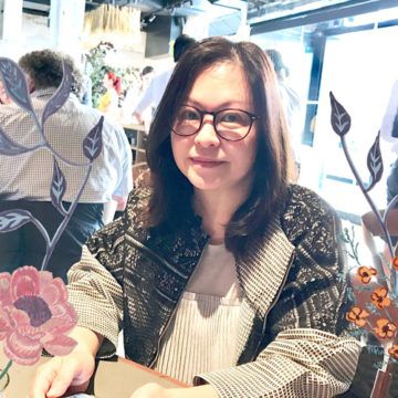 A portrait of Maggie Lam seated at a table in a restaurant. She has shoulder length straight brown hair and dark framed glasses. The photo is overlayed with some hand drawn floral motifs.