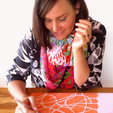 A tightly cropped photo of Marni Stuart working on an orange and pink abstract painting at a wooden table. She is wearing a black and white abstract print long sleeve top and a colourful floral design scarf. Marni has shoulder length straight brown hair.