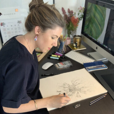 Laura of MONBER and B is shown seated at a desk sketching on a drawing pad. There is a reference image of a leaf shown on her computer screen. Laura has dark blonde hair and is wearing a short sleeved black shirt and colourful teardrop shaped earrings.