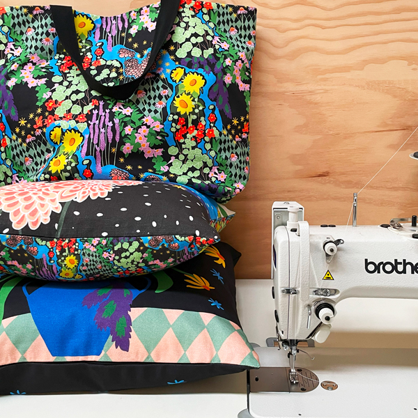 Three custom made products, manufactured by Next State, are stacked to the left of an industrial sewing machine. There is a 50cm square cushion on the bottom, a 45cm square cushion on that, and a tote bag on top.