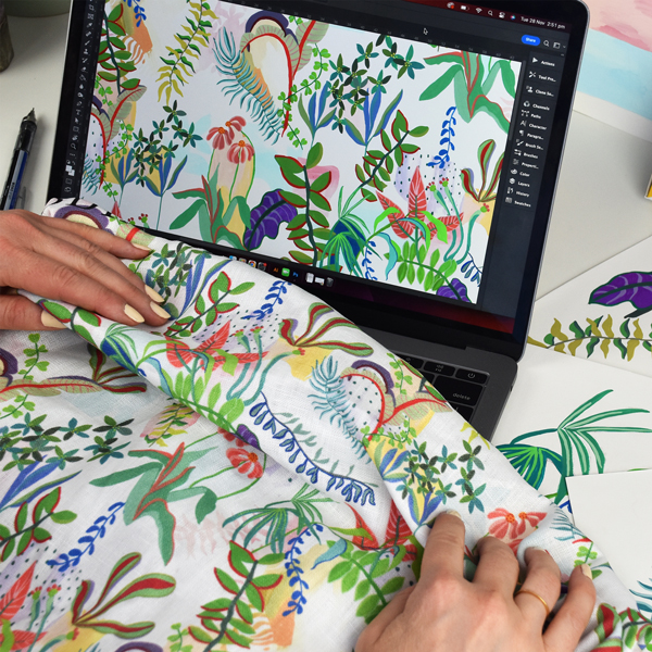 Tightly cropped photo of hands holding custom printed fabric in front of a laptop showing the digital design of the fabric print.