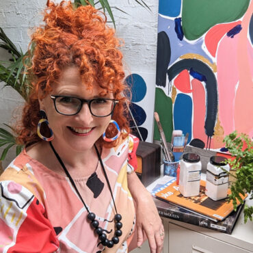 Ellen Mckenna is a white woman with bright orange-red, curly hair that is loosely tied back. She is wearing a short sleeve top with a bold, colourful, abstract print. Ellen is sitting beside an array of art supplies and paintings sitting atop a bench.