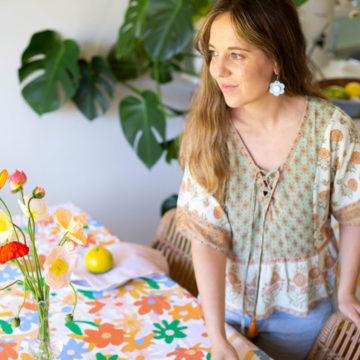 A woman with long wavy blonde hair wearing a bohemian style blouse and daisy shaped blue earrings is sitting at a table looking off to the left. The table is dressed with a printed tablecloth designed by Linen Jungle featuring a multi-coloured floral pattern.