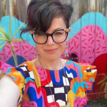 Bronwyn Seedeen is pictured wearing a bright and bold short sleeved shirt standing in front of a colourfully painted wooden fence. She has short dark hair and is wearing black framed glasses with gold accents.