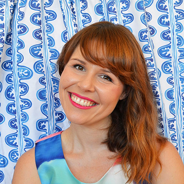 A portrait of Lauren Malone of The Indigo Room posed in front of her Clam Stripe in Ocean fabric. Lauren has shoulder length wavy red hair with a fringe and is wearing a blue, turquoise, white and coral coloured tanktop.