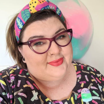A close cropped photo of Natalie Perkins of Fancy Lady Industries, a white woman with her short hair held back with colourful headband. She is wearing a patterned black shirt with small colourful rectangles and dark-magenta cats eye glasses.