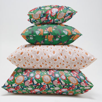 A stack of four cushions made from prints of Vanessa Holliday's designs is viewed from side-on. The top and bottom cushion are made from fabric that features dragonfruit and Sturt's Desert Pea, the second from the top shows a green, pink and orange floral design, and the third cushion from the top shows an abstract beige print with dots and lines.