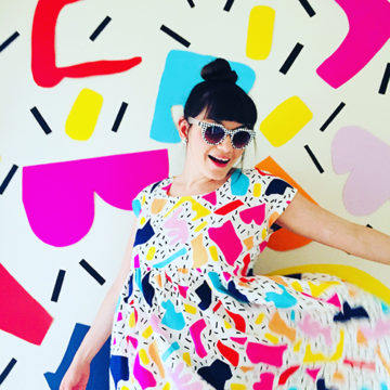 A white woman with dark hair tied up in a bun stands in front of a white background with bright abstract shapes. She is wearing black and white sunglasses and a dress that is made using the same Whimsy Kaleidoscope design that is shown in the background.