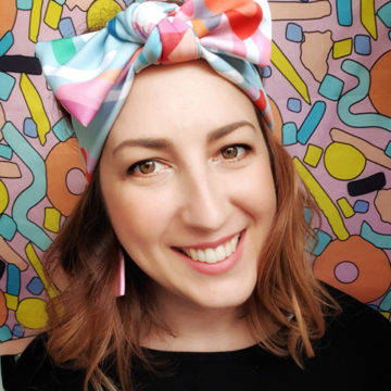 A tightly cropped portrait of a smiling white woman with brown eyes and shoulder length, wavy, red hair held back with a headband tied in a bow on top of her head. The headband fabric is printed with an abstract pastel design. The background of the portrait is another design by Winter Bloom Designs that features brightly coloured organic shapes on a pink background.