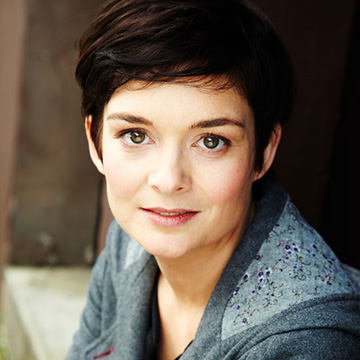 A headshot of Mel Armstrong, a white woman with pixie cut dark brown hair and brown eyes. She is wearing a mid-grey wool hooded jacket.