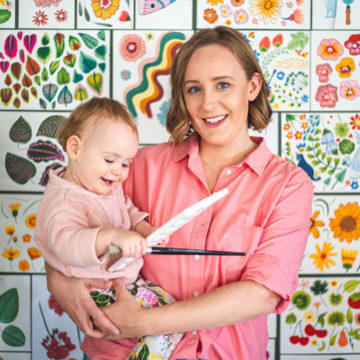 Megan Isabella is a white woman with chin-length light brown hair. She is wearing a coral-pink button up shirt and is holding a baby wearing a light pink jumper and colourful patterned pants. They are posed in front of a wall decorated with a tightly grouped collection of Megan's own artwork.