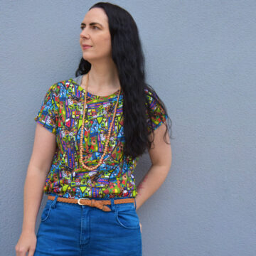 Ang Watson of Antayjo Art is standing in front of a blue wall looking off to the left. She is wearing a short sleeved shirt printed in one of her vibrant designs with blue jeans, a brown belt, and a beaded necklace. She has long dark brown wavy hair.